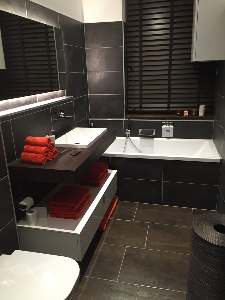 Cette photo montre une petite salle de bain principale moderne avec une baignoire encastrée, WC suspendus, un carrelage noir, un mur marron, un lavabo encastré et un plan de toilette en bois.