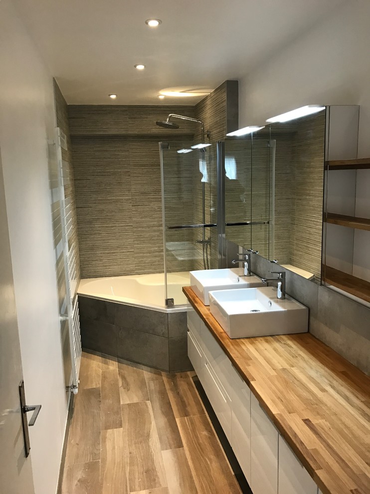 Bild på ett litet funkis en-suite badrum, med ett hörnbadkar, grå kakel, grå väggar, ett nedsänkt handfat, träbänkskiva och brunt golv