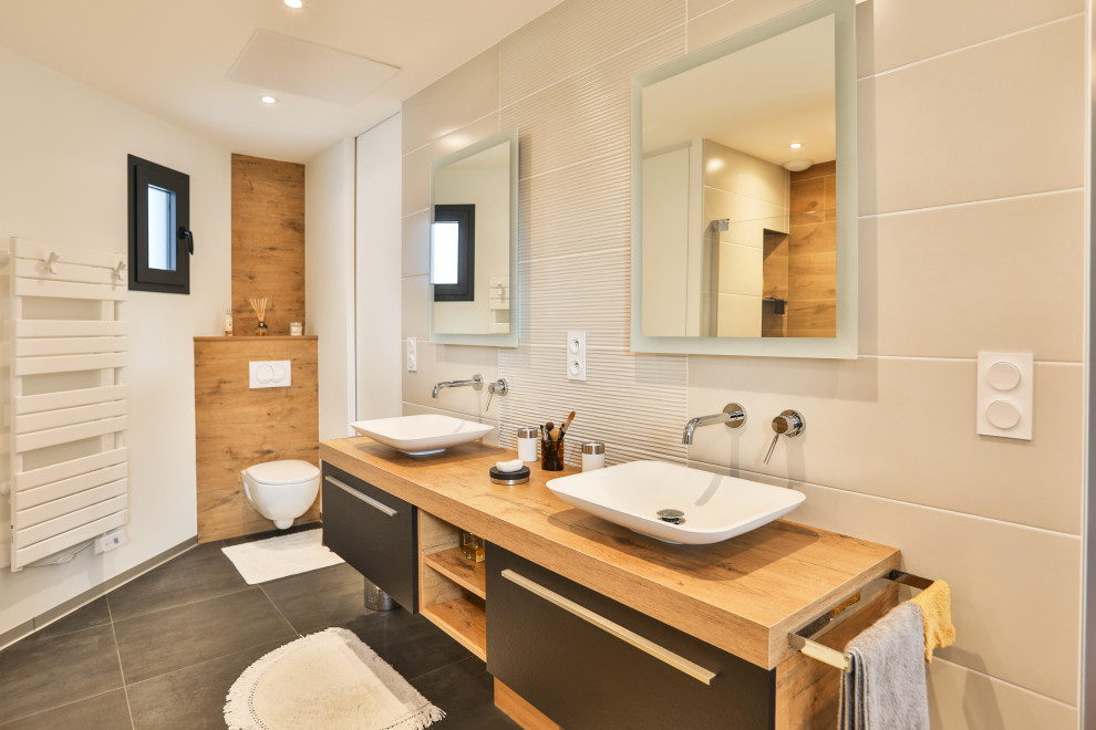 Cette image montre une salle de bain design avec un placard à porte plane, des portes de placard grises, WC suspendus, un carrelage beige, un mur blanc, une vasque, un plan de toilette en bois, un sol gris, un plan de toilette marron, meuble double vasque et meuble-lavabo suspendu.