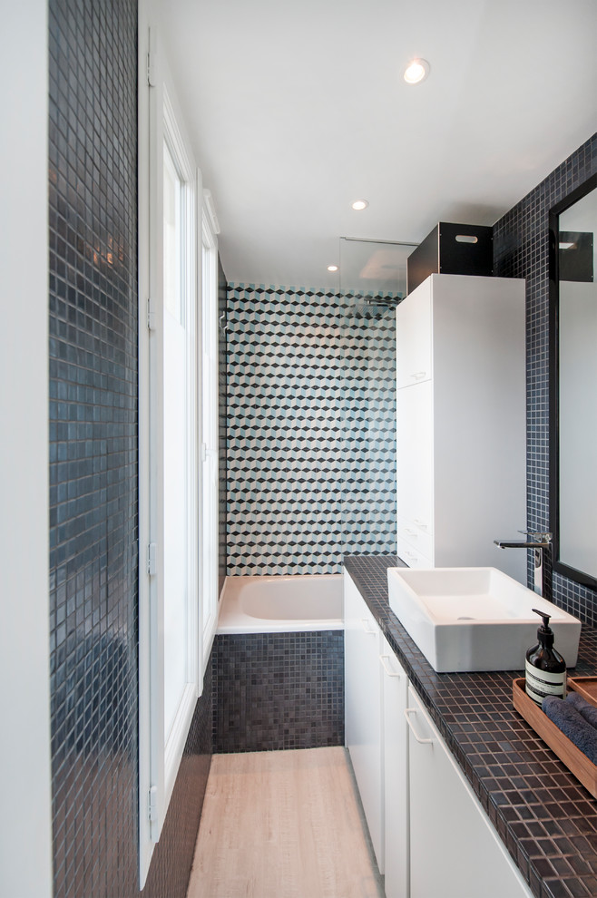 На фото: узкая и длинная ванная комната в современном стиле с плоскими фасадами, белыми фасадами, накладной ванной, душем над ванной, разноцветной плиткой, настольной раковиной, столешницей из плитки, бежевым полом и черной столешницей с