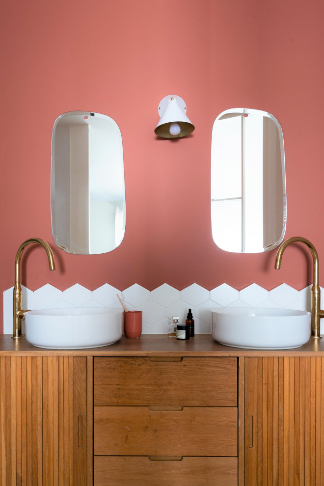 Imagen de cuarto de baño tropical con puertas de armario de madera oscura, paredes rosas, lavabo sobreencimera, encimera de madera, baldosas y/o azulejos blancos, espejo con luz y armarios con paneles lisos