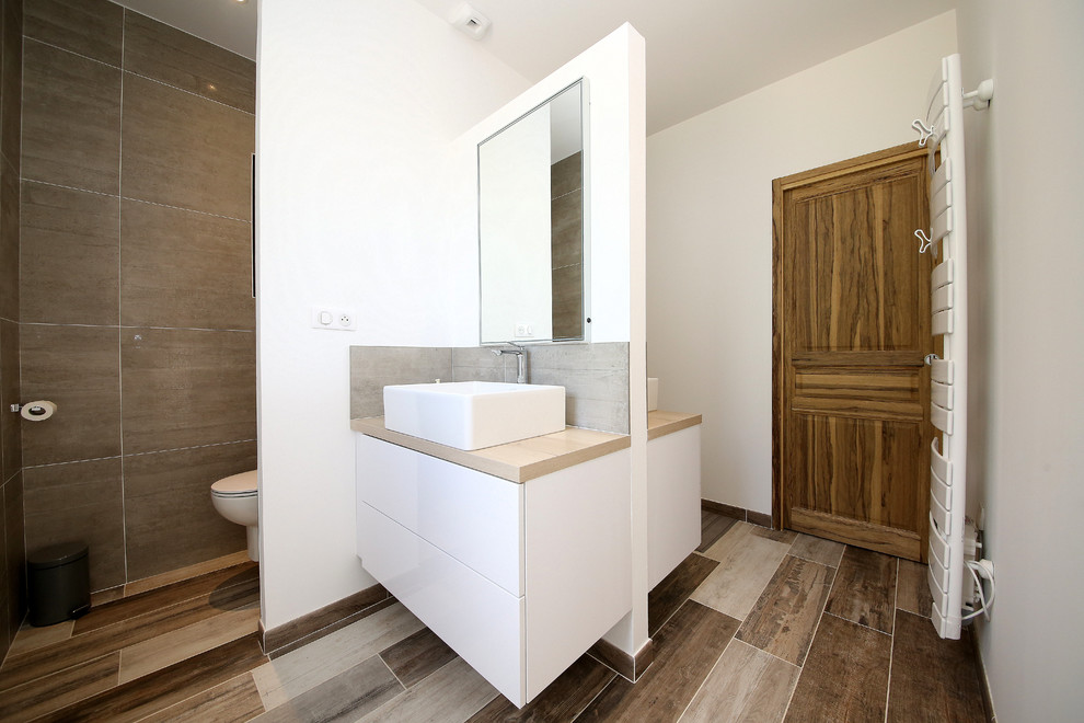Foto på ett medelhavsstil badrum med dusch, med luckor med profilerade fronter, en vägghängd toalettstol, grå kakel, vita väggar, ett konsol handfat, träbänkskiva och brunt golv