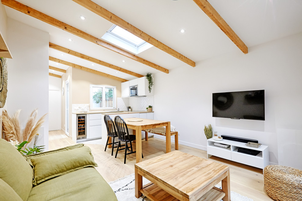 Imagen de sala de estar abierta escandinava de tamaño medio sin chimenea con paredes blancas, suelo de madera clara, televisor colgado en la pared, suelo beige y vigas vistas