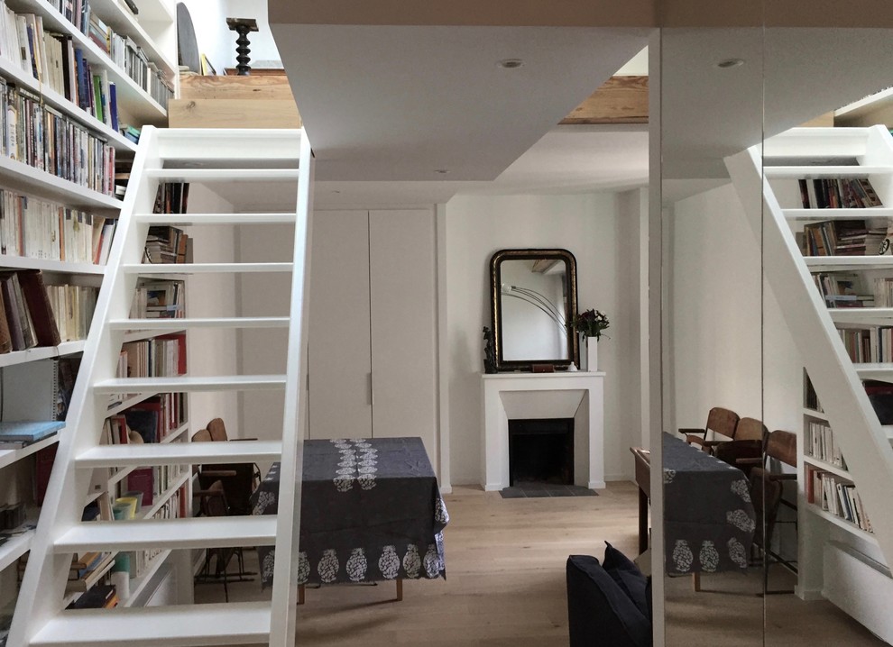Cette image montre une salle de séjour design de taille moyenne et ouverte avec un mur blanc, parquet clair, une bibliothèque ou un coin lecture, un sol marron, une cheminée standard et un manteau de cheminée en plâtre.