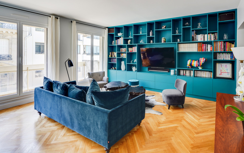 На фото: большая открытая гостиная комната в современном стиле с с книжными шкафами и полками, синими стенами, светлым паркетным полом, телевизором на стене и бежевым полом без камина с