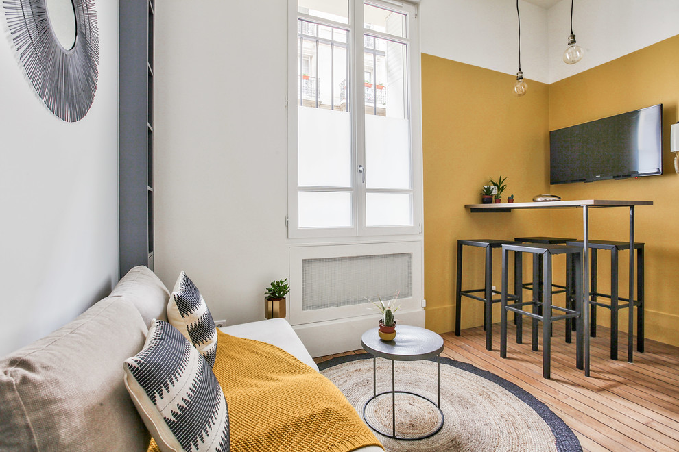 Cette image montre une salle de séjour urbaine de taille moyenne et ouverte avec un mur jaune, un sol en bois brun, aucune cheminée, un téléviseur fixé au mur, un sol marron et une bibliothèque ou un coin lecture.
