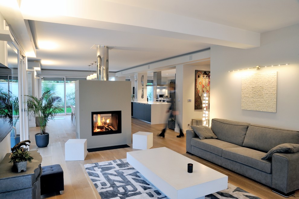 Cette image montre une grande salle de séjour design ouverte avec un mur gris, parquet clair, une cheminée double-face, un manteau de cheminée en béton, un sol beige et un téléviseur fixé au mur.