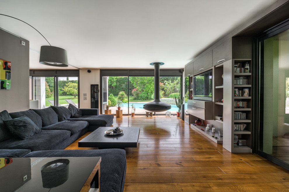 Cette image montre un salon design avec cheminée suspendue, un mur gris, un sol en bois brun, un manteau de cheminée en métal, un téléviseur encastré, un sol marron et éclairage.