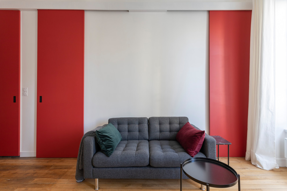 Modern inredning av ett litet allrum med öppen planlösning, med röda väggar, ljust trägolv, en standard öppen spis, en spiselkrans i trä, en dold TV och beiget golv