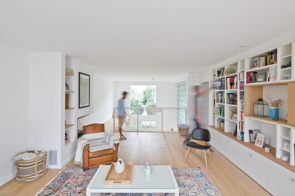 Diseño de biblioteca en casa abierta contemporánea de tamaño medio con paredes blancas, suelo de madera clara, chimenea de esquina, marco de chimenea de yeso y televisor retractable