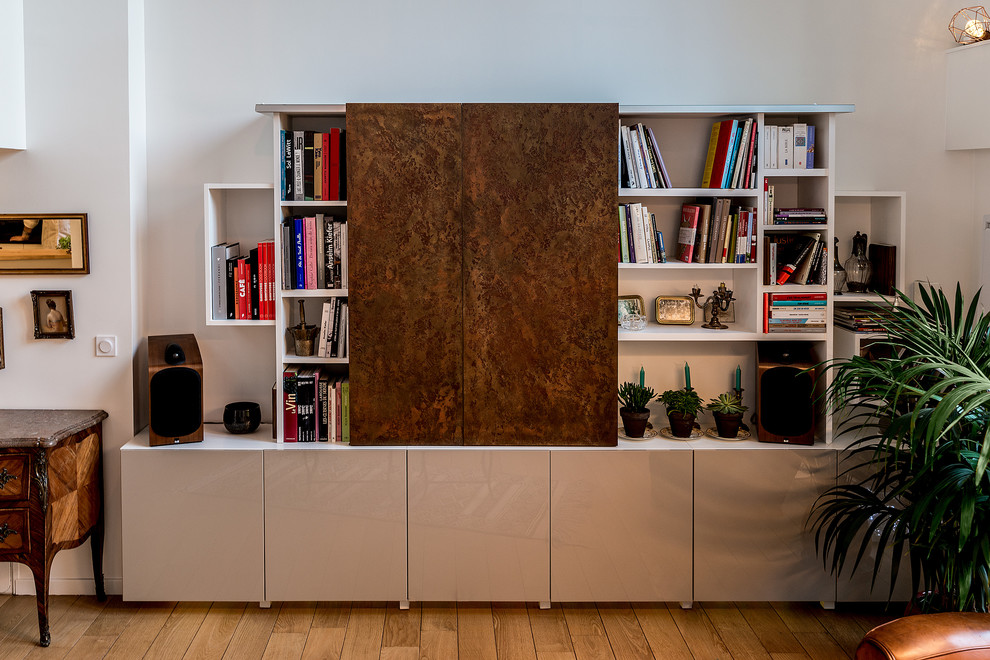 Modelo de biblioteca en casa abierta contemporánea de tamaño medio con paredes blancas, suelo de madera clara y televisor retractable