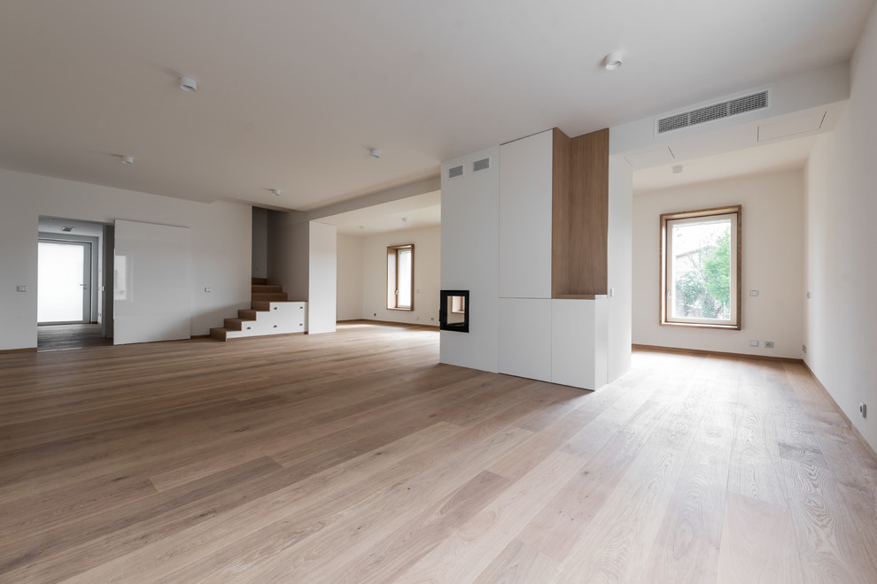 Foto de salón abierto nórdico grande con paredes blancas, suelo de madera clara, chimenea de doble cara, marco de chimenea de yeso y suelo marrón