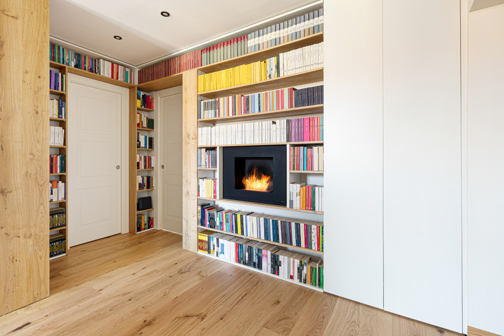 Imagen de biblioteca en casa abierta escandinava grande con paredes blancas, suelo de madera clara, chimeneas suspendidas, marco de chimenea de metal, televisor colgado en la pared y bandeja