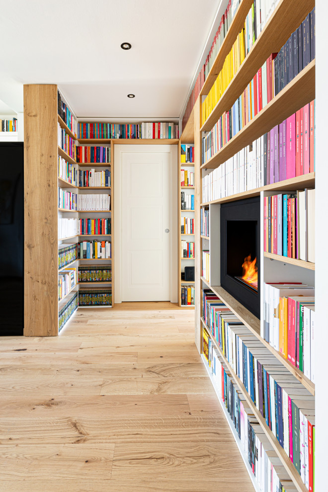 Modelo de biblioteca en casa abierta escandinava grande con paredes blancas, suelo de madera clara, chimeneas suspendidas, marco de chimenea de metal, televisor colgado en la pared y bandeja