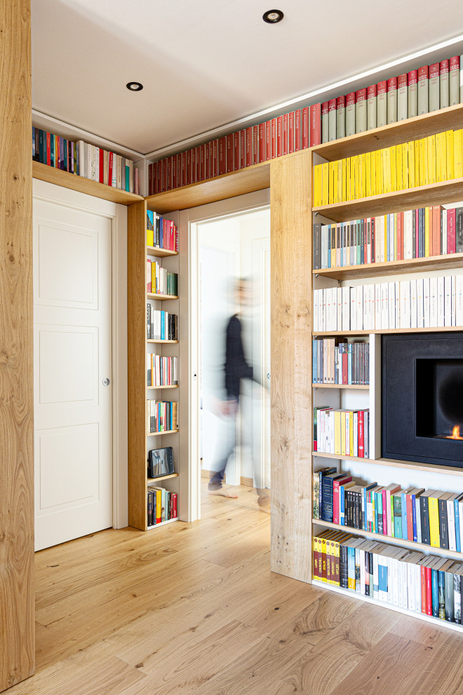 Bild på ett stort nordiskt allrum med öppen planlösning, med ett bibliotek, vita väggar, ljust trägolv, en hängande öppen spis, en spiselkrans i metall och en väggmonterad TV