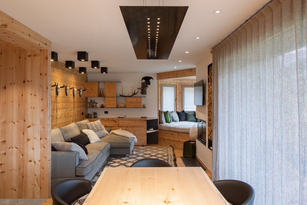 Imagen de sala de estar abierta rústica con paredes blancas, suelo de madera clara, estufa de leña y televisor colgado en la pared