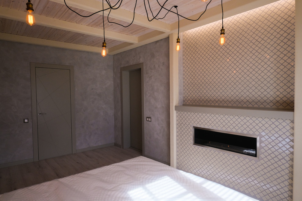Diseño de dormitorio principal industrial de tamaño medio con paredes grises, suelo laminado, chimenea lineal, marco de chimenea de yeso y suelo gris