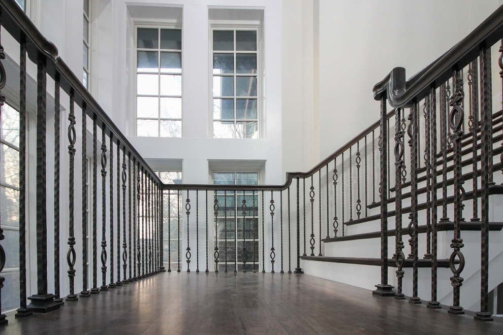 Пример оригинального дизайна: большая деревянная лестница на больцах в стиле неоклассика (современная классика) с деревянными ступенями и стеклянными перилами