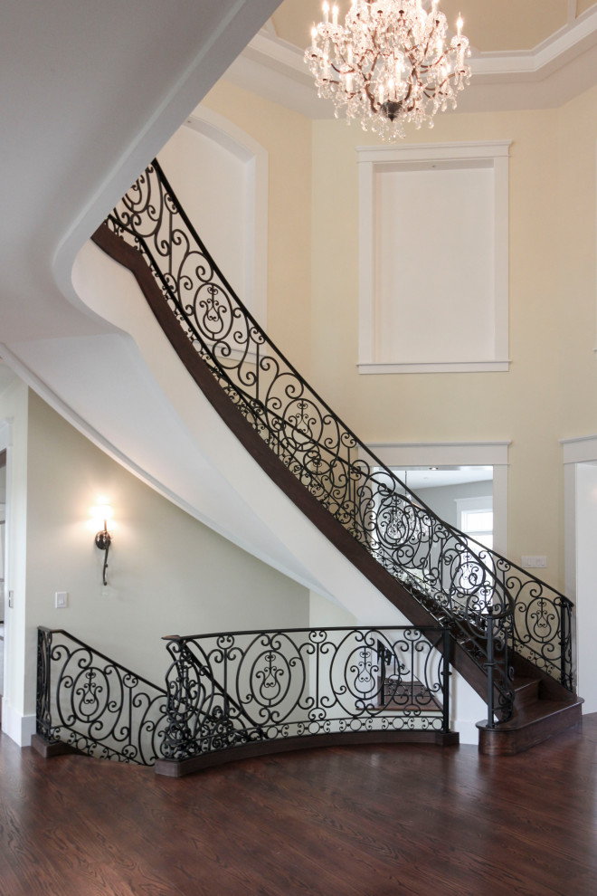Стильный дизайн: огромная изогнутая деревянная лестница в стиле неоклассика (современная классика) с деревянными ступенями и металлическими перилами - последний тренд