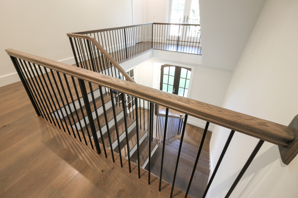 Пример оригинального дизайна: большая угловая лестница в стиле неоклассика (современная классика) с деревянными ступенями, крашенными деревянными подступенками, металлическими перилами и панелями на части стены