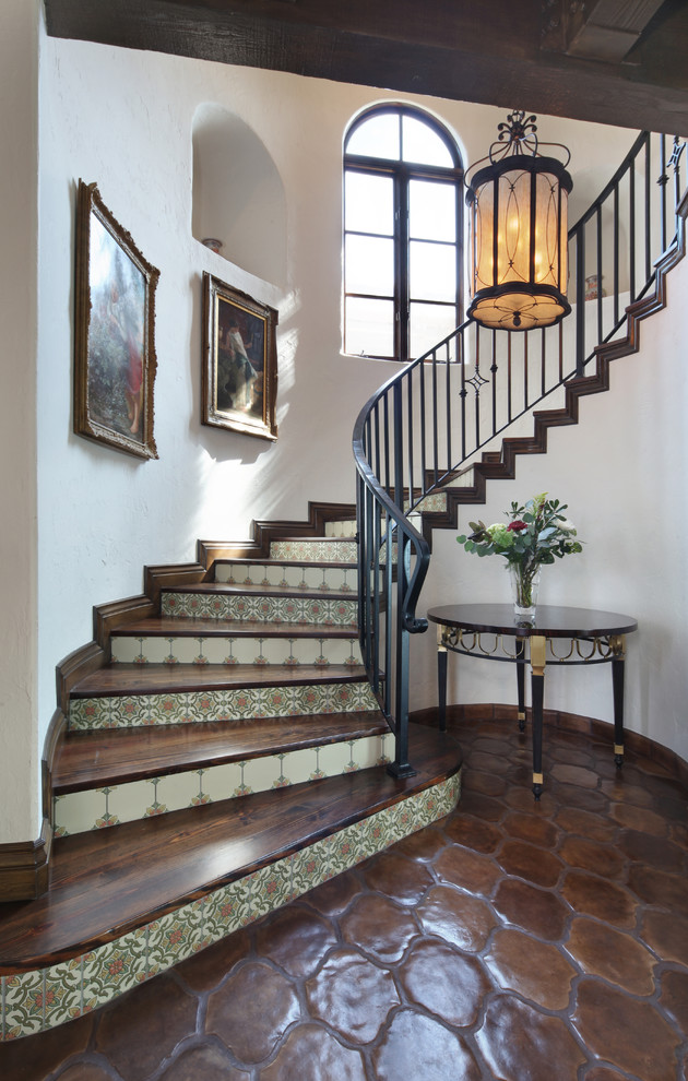 На фото: большая изогнутая лестница в средиземноморском стиле с деревянными ступенями и подступенками из плитки с