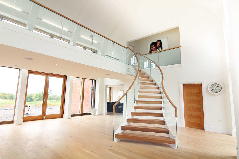 Trendy wooden curved glass railing staircase photo in West Midlands with wooden risers