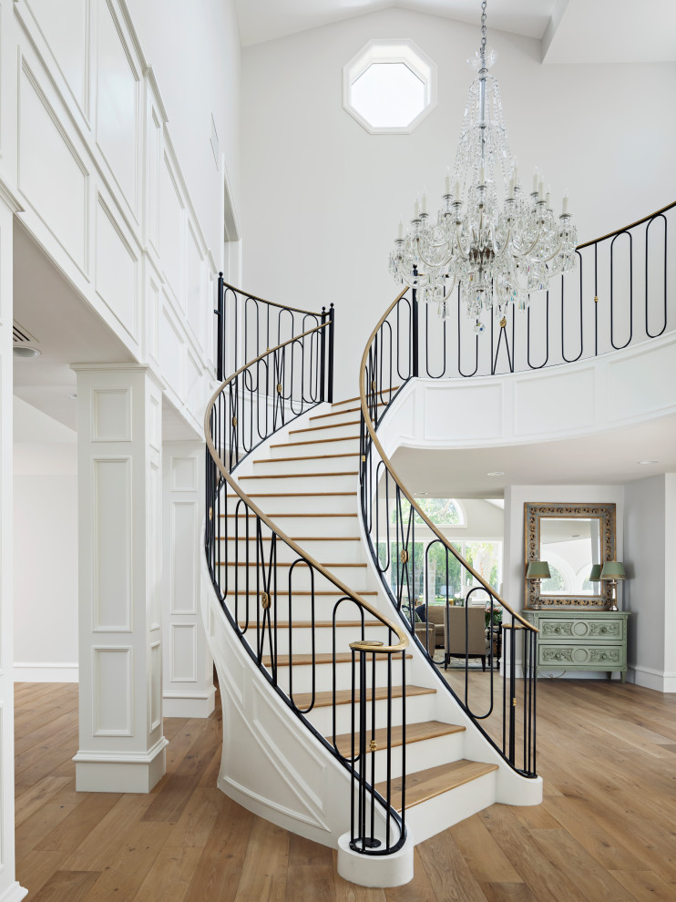 На фото: огромная изогнутая лестница в классическом стиле с деревянными ступенями, металлическими перилами и крашенными деревянными подступенками с