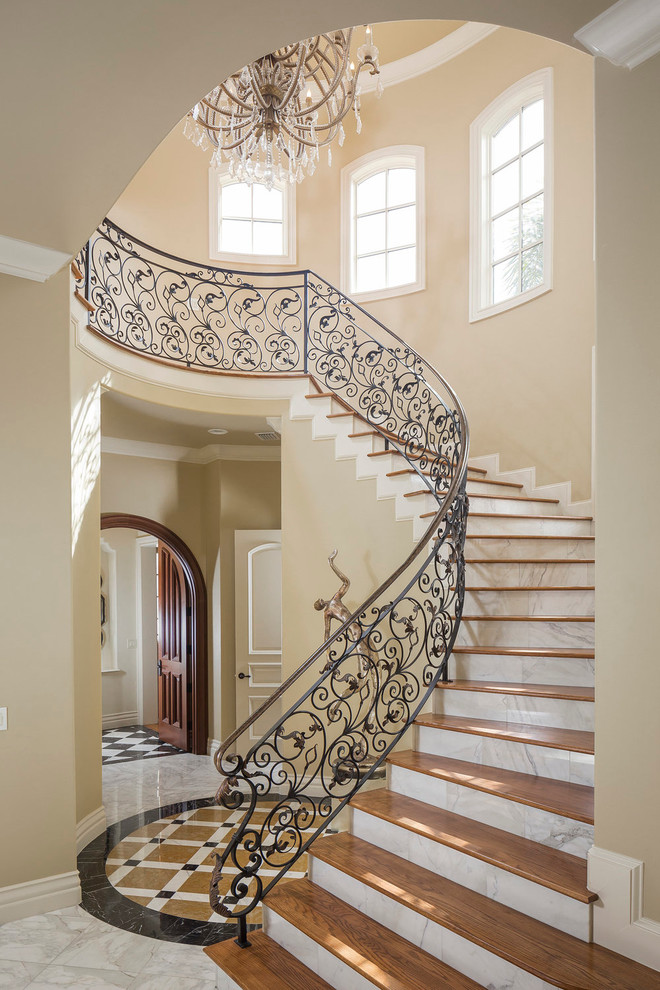 На фото: большая изогнутая лестница в средиземноморском стиле с деревянными ступенями, металлическими перилами и подступенками из мрамора