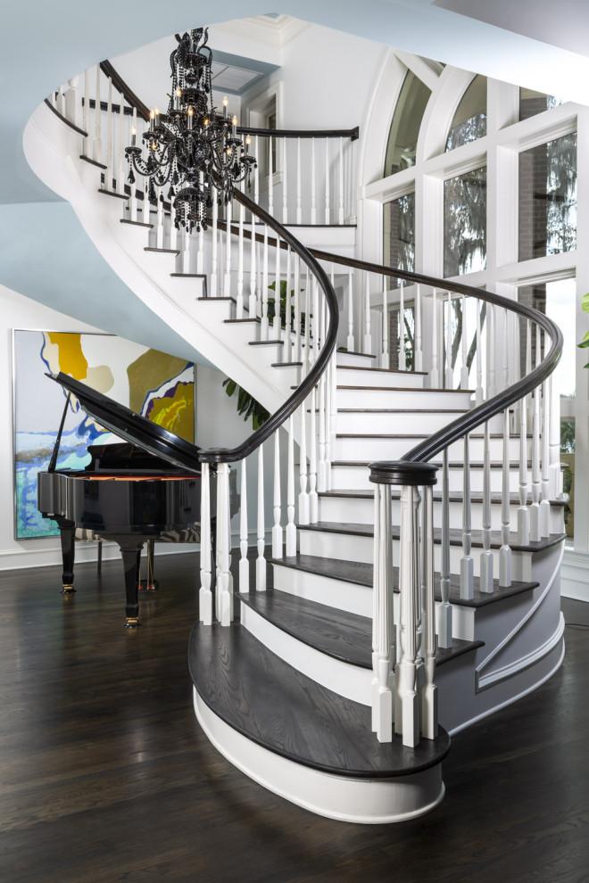 Идея дизайна: огромная изогнутая лестница в стиле неоклассика (современная классика) с деревянными ступенями, крашенными деревянными подступенками и деревянными перилами