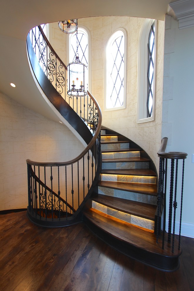 На фото: огромная изогнутая лестница в классическом стиле с деревянными ступенями, подступенками из сланца и деревянными перилами с