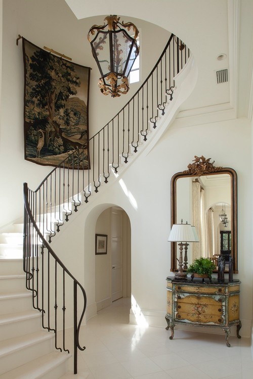 foyer  with white tiled floor and curved iron staircase and large tapestry hanging on staircase wall