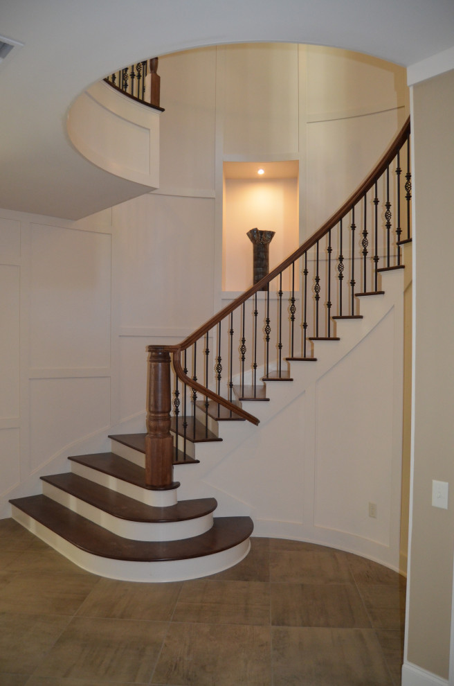 На фото: большая изогнутая лестница в стиле неоклассика (современная классика) с деревянными ступенями, крашенными деревянными подступенками, металлическими перилами и панелями на части стены