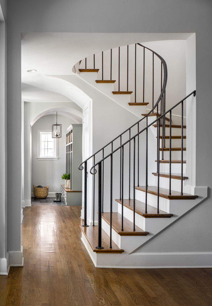 Пример оригинального дизайна: большая изогнутая лестница в стиле неоклассика (современная классика) с деревянными ступенями, крашенными деревянными подступенками и металлическими перилами