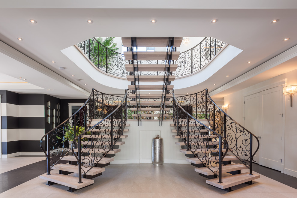 Пример оригинального дизайна: большая изогнутая лестница в стиле неоклассика (современная классика) с ступенями из известняка и металлическими перилами без подступенок
