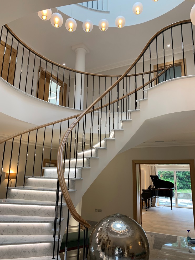 На фото: большая изогнутая лестница в стиле фьюжн с мраморными ступенями, подступенками из мрамора и металлическими перилами с