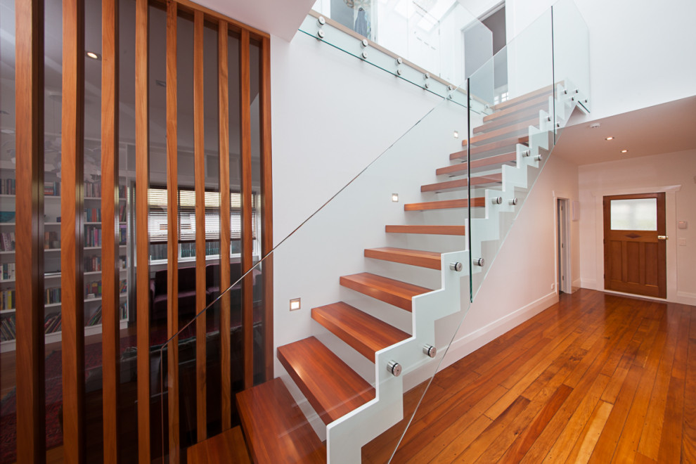 Стильный дизайн: большая лестница на больцах в современном стиле с деревянными ступенями и стеклянными перилами без подступенок - последний тренд