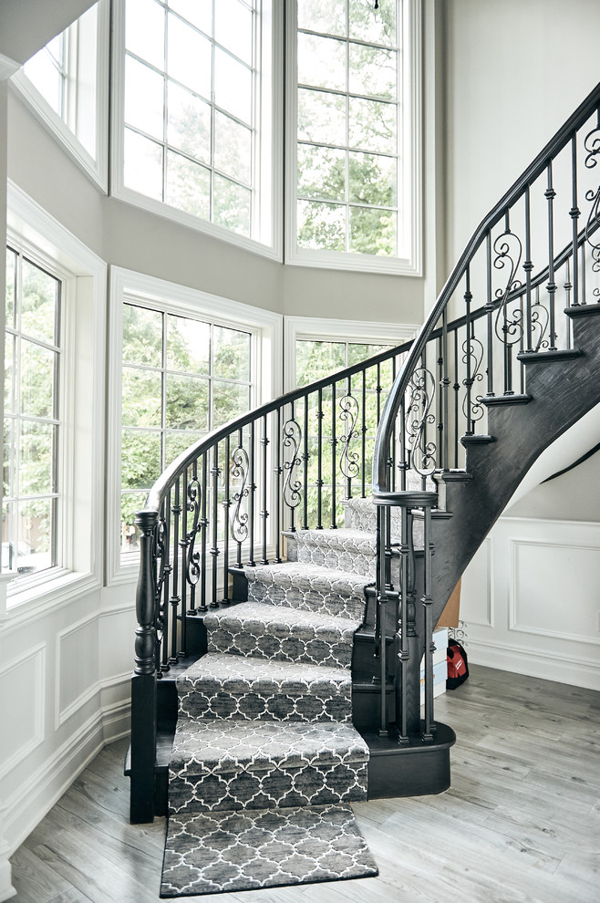 Стильный дизайн: большая изогнутая лестница в стиле неоклассика (современная классика) с ступенями с ковровым покрытием, ковровыми подступенками и металлическими перилами - последний тренд