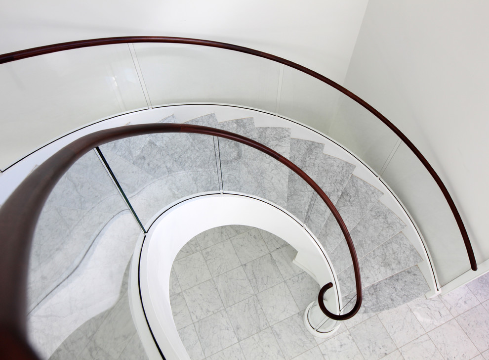 Cette photo montre un très grand escalier courbe tendance en marbre avec des contremarches en marbre et un garde-corps en verre.