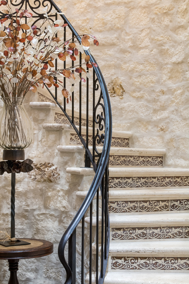 Aménagement d'un grand escalier courbe méditerranéen avec des marches en pierre calcaire, des contremarches carrelées et un garde-corps en métal.