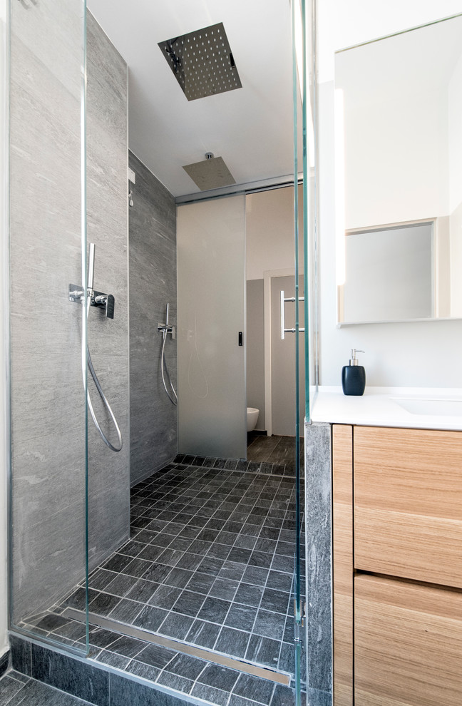 Idéer för att renovera ett mellanstort funkis badrum med dusch, med släta luckor, skåp i ljust trä, en dubbeldusch, en vägghängd toalettstol, grå kakel, porslinskakel, klinkergolv i porslin, grå väggar, ett undermonterad handfat och bänkskiva i akrylsten
