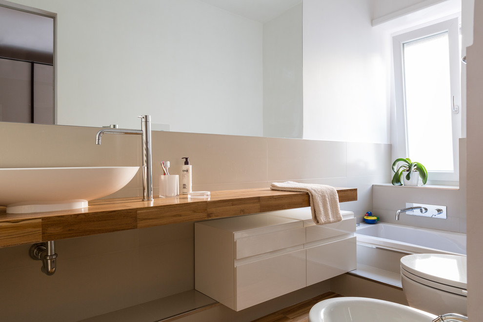 Foto på ett funkis badrum med dusch, med släta luckor, vita skåp, ett platsbyggt badkar, en bidé, beige kakel, beige väggar, mellanmörkt trägolv, ett fristående handfat, träbänkskiva och beiget golv