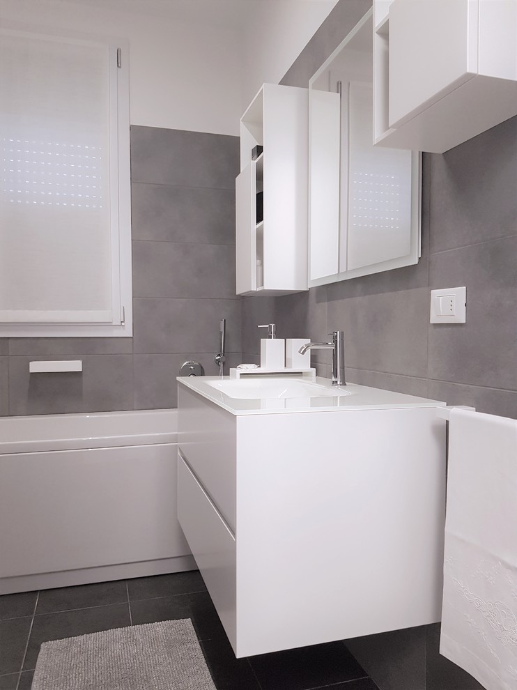 Photo of a medium sized modern ensuite bathroom in Milan with flat-panel cabinets, white cabinets, a freestanding bath, grey tiles, porcelain tiles, white walls, ceramic flooring, an integrated sink, glass worktops, grey floors and white worktops.