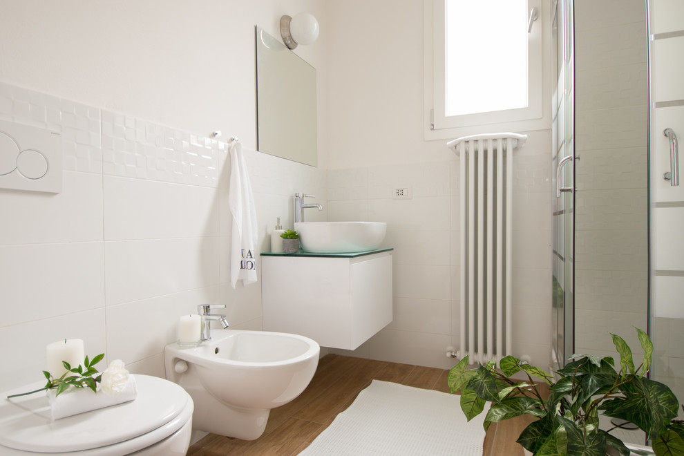 На фото: ванная комната среднего размера в современном стиле с плоскими фасадами, биде, белой плиткой, белыми стенами, светлым паркетным полом, настольной раковиной и стеклянной столешницей