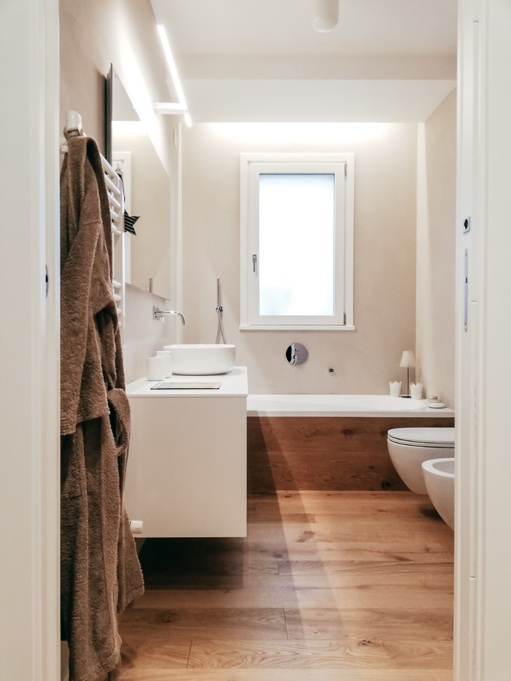Mittelgroßes Modernes Duschbad mit beiger Wandfarbe, hellem Holzboden, weißen Schränken, Badewanne in Nische, Wandtoilette, Aufsatzwaschbecken, beigem Boden und weißer Waschtischplatte in Mailand
