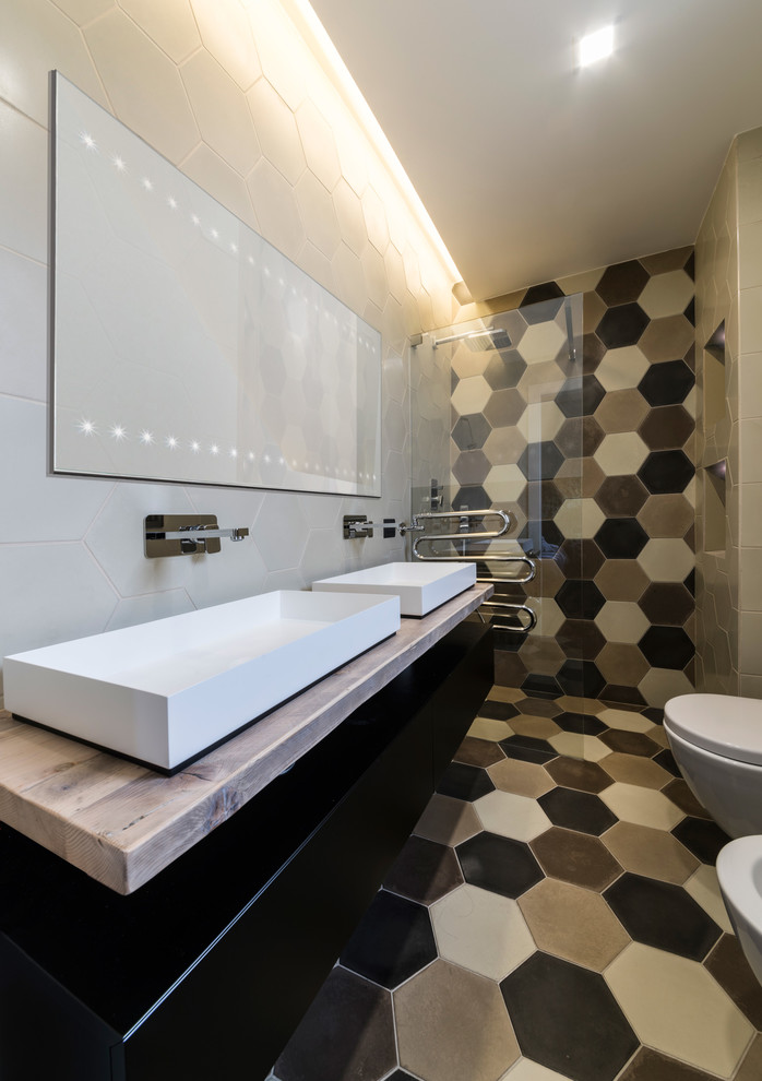 Modern inredning av ett mellanstort badrum med dusch, med svarta skåp, en kantlös dusch, en vägghängd toalettstol, beige kakel, vit kakel, svart kakel, cementkakel, beige väggar, cementgolv, ett avlångt handfat, träbänkskiva och släta luckor