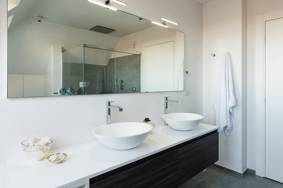 Medium sized modern ensuite bathroom in Milan with recessed-panel cabinets, grey cabinets, a hot tub, a built-in shower, a vessel sink, solid surface worktops, a sliding door, white worktops, double sinks, a freestanding vanity unit, a wall mounted toilet, white walls, ceramic flooring and grey floors.