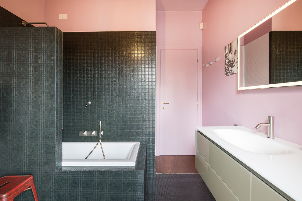 Inspiration for a medium sized contemporary ensuite bathroom in Milan with a built-in bath, a wall mounted toilet, black tiles, mosaic tiles, pink walls, an integrated sink, glass worktops, green floors and white worktops.