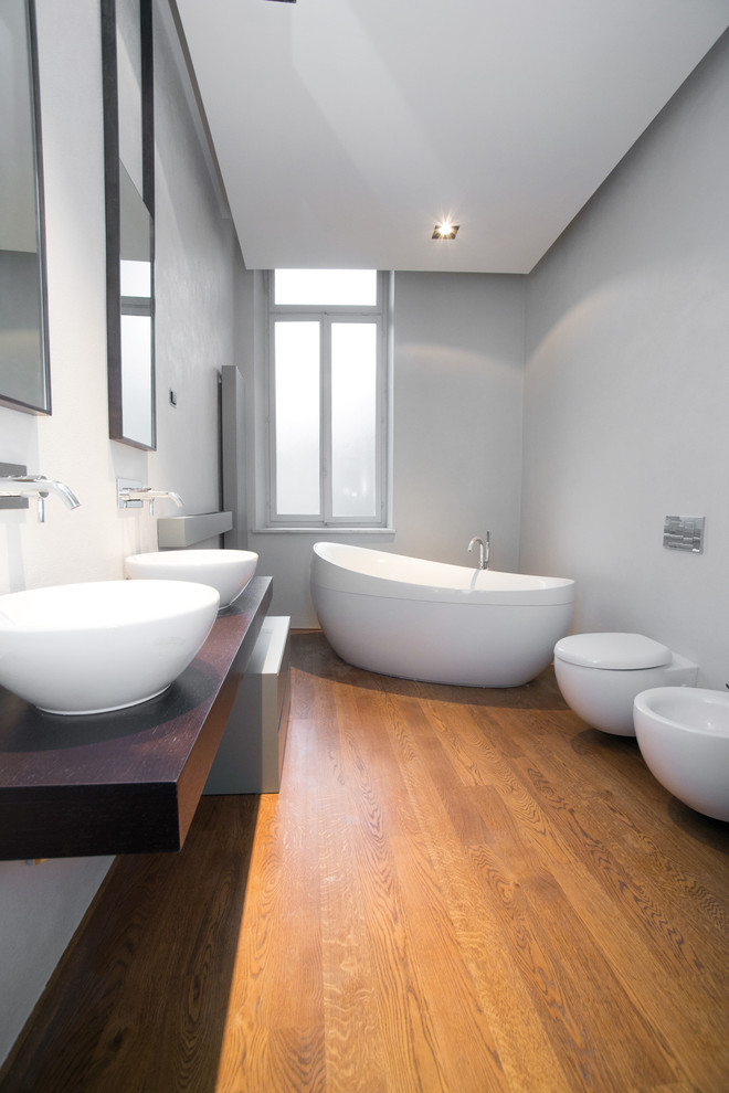 Idées déco pour une grande salle de bain principale contemporaine avec une baignoire indépendante, WC suspendus, un mur gris, un sol en bois brun, une vasque, un plan de toilette en bois et un plan de toilette marron.