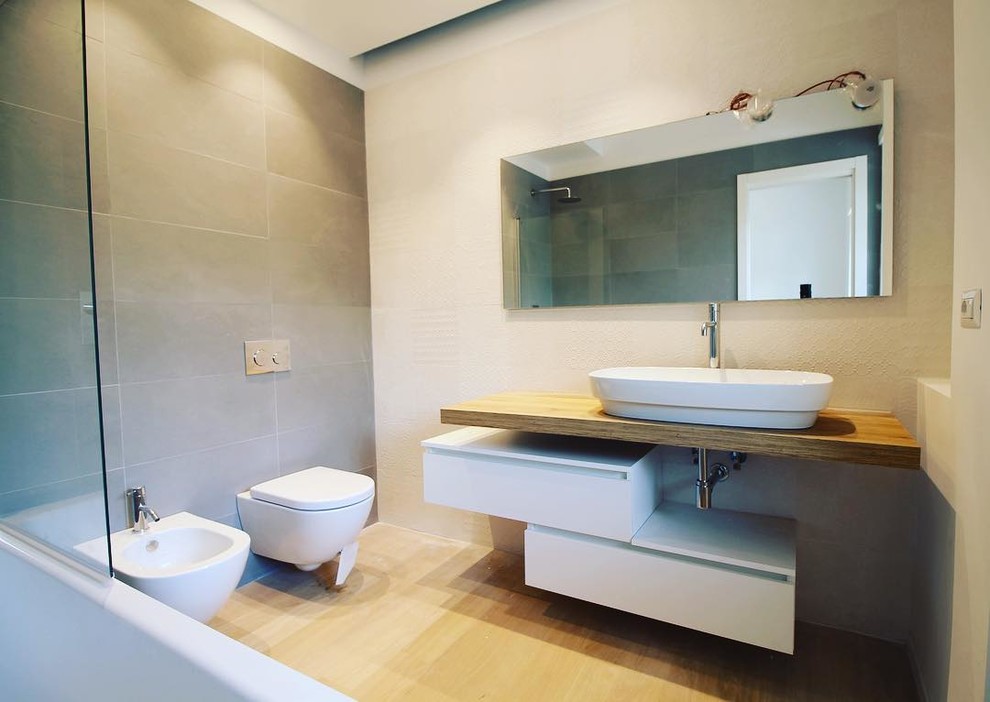 Aménagement d'une salle de bain principale moderne en bois clair avec un placard à porte plane, une baignoire d'angle, un combiné douche/baignoire, un carrelage gris, des carreaux de porcelaine, un mur blanc, parquet clair, une vasque, un plan de toilette en bois et une cabine de douche à porte battante.