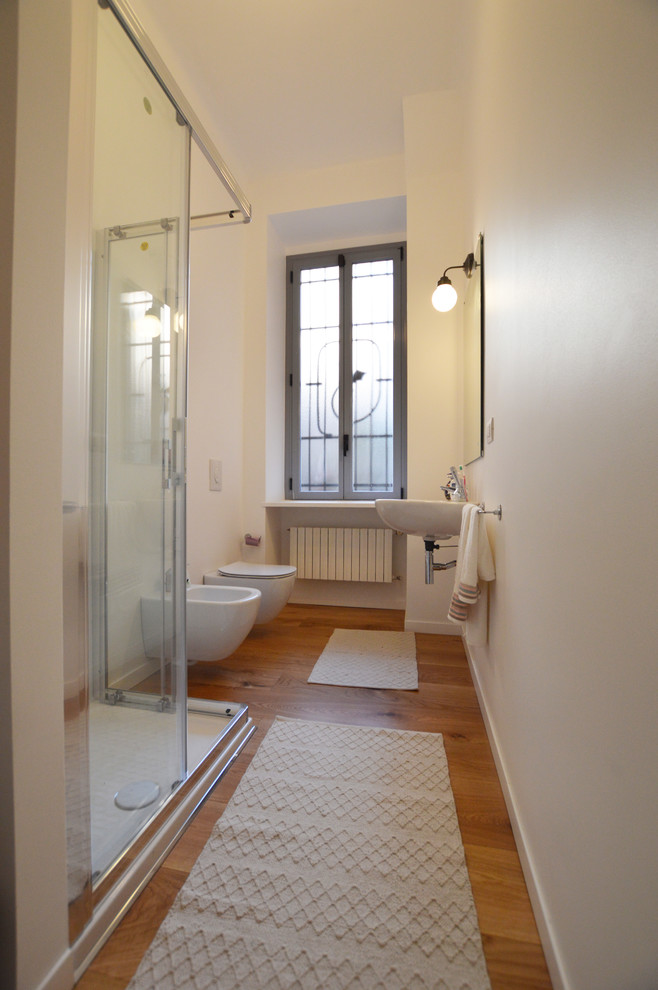 Modern inredning av ett mellanstort badrum med dusch, med en hörndusch, en toalettstol med separat cisternkåpa, vita väggar, ljust trägolv, ett väggmonterat handfat, brunt golv och dusch med skjutdörr